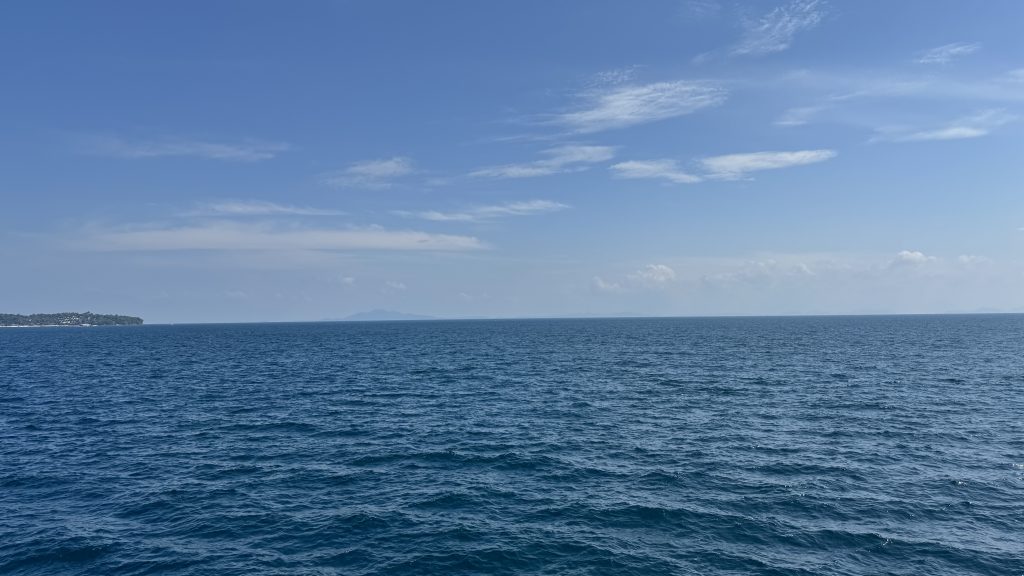 A wide-angle shot of the horizon over a deep blue ocean under a bright, clear sky with soft white clouds.