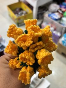 A hand holds a vibrant bouquet of crocheted yellow flowers, featuring multiple bell-shaped petals and green detailing at the base. 