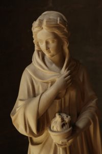 This vertical close-up photo features a classical-style sculpture of a woman carved from a light-colored, smooth stone. She is depicted with a serene, downcast expression, holding a small decorative bowl or vessel in her hands against a dark background.