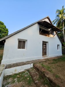A white traditional building with a sloped roof, wooden door, and a small window sits in a sunny setting, with lush greenery and old stone steps, at Niramkaitha Kotta Temple, Vallikunnu North, Malappuram.