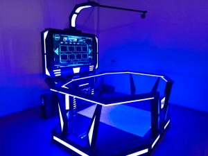 A sleek VR gaming setup with bright blue LED lights, a large central screen, a hexagonal frame for movement, and an overhead sensor arm, all placed indoors against a plain wall.
