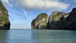 Calm ocean bay with boats near green cliffs and rocky islands under a bright sky.