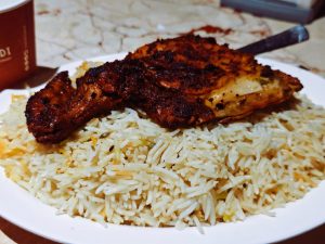 A plate of aromatic, fluffy white rice topped with a well-cooked, char-grilled chicken piece, showing a crispy, darkened exterior and juicy interior. 