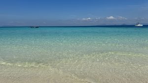 Wide tropical beach with clear shallow water, a small boat, and a catamaran under a blue sky.
