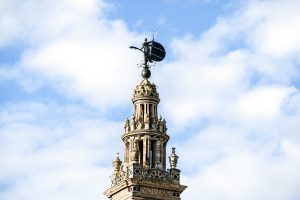 La Giralda, a tall, ornate tower rises against a backdrop of softly scattered clouds. The pinnacle features a golden dome topped with El Giraldillo, a large 16th century bronze statue representing Faith