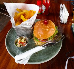 Beef burger with lettuce, tomato, onion, and a cherry, served with potato wedges and fruit salsa on a wooden restaurant table.