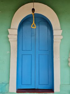 A bright blue wooden door set inside a white arch on a green wall, with a hanging handle above. A colorful and balanced architectural detail of the Mishkal Mosque in Kuttichira, Kozhikode.