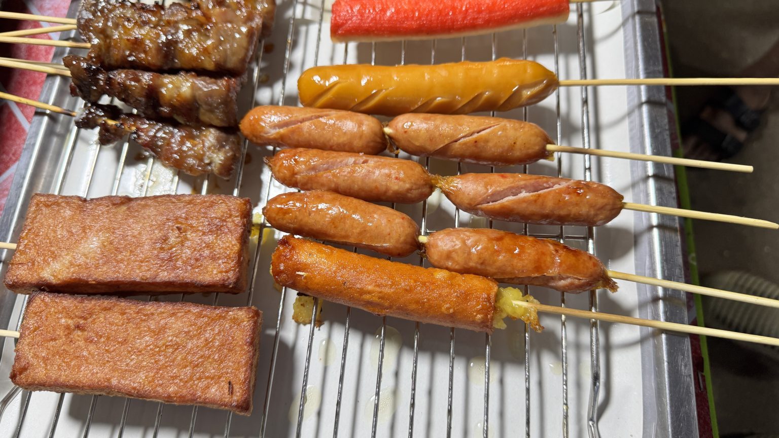 Close-up of grilled snacks on wooden skewers over a metal tray: fried tofu or meat, small cut-marked sausages, dark grilled meat, and a long sausage stuffed with corn.