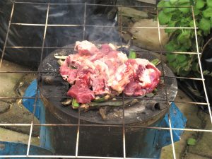 Raw meat grills over an open flame on a round metal grate, surrounded by fresh greens in an outdoor setting with smoke rising into the air.