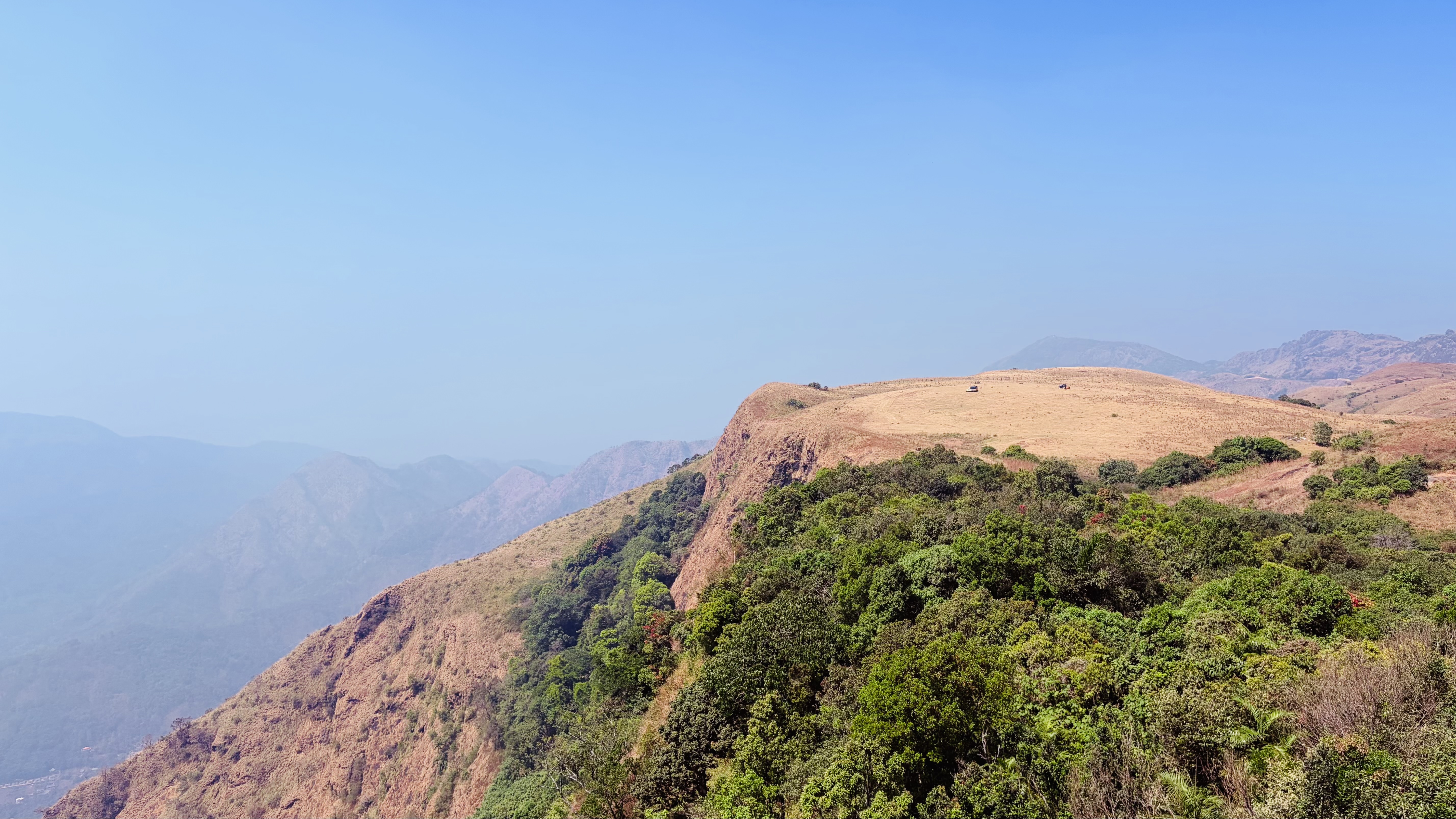 A panoramic view of a rugged landscape featuring a steep, grassy hilltop that transitions into a rocky cliff.