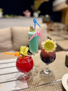 Three vibrant tropical cocktails in various glasses - one red, one bright blue, and one deep purple, garnished with citrus slices and colorful bendy straws. The drinks are arranged on a woven placemat at an outdoor lounge at night.