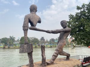 Sculpture of two figures by a riverside, one holding a briefcase with a missing midsection. Another, straining to hold them together, under a cloudy sky.