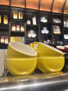 Four yellow ceramic cups are stacked on a metal counter in a cafe, with shelves of coffee products and mugs blurred in the background.