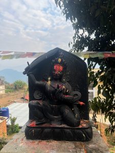 A black stone statue of the deity Manjushri is perched on a stone wall overlooking a valley. The figure is depicted in a kneeling warrior pose, holding a raised sword in the right hand and a lotus stem in the left. The statue's face and body are decorated with bright red and yellow ritual powders. Colorful prayer flags and green trees frame the bac