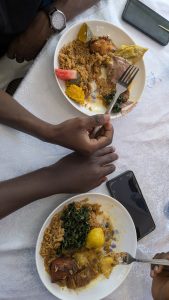 Top-down view of two plates of food on a white tablecloth with rice, vegetables, plantains, beans, pumpkin, and meat, as two people prepare to eat using a fork and spoon, with two smartphones on the table