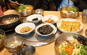 A top-down view of a wooden table filled with various Korean dishes. Large brass bowls contain bibimbap with fresh vegetables and rice, while smaller side dishes of kimchi, seaweed, rolled omelets, and marinated beef are arranged in the center.