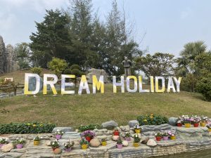 A big “Dream Holiday” sign stands on a grassy hill with colorful flowers below. Trees surround the area, and a stone statue nearby makes the place feel cheerful and welcoming.
