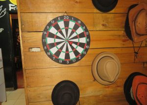 A dartboard with a single red dart stuck in the bullseye is mounted on a rustic wooden wall made of horizontal planks. Surrounding the dartboard are several hats of different styles and colors, including fedoras and cowboy hats in shades of brown, black, and orange.