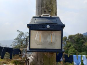 Solar-powered outdoor wall lamp with two bulbs mounted on a wooden pole, with trees, hills, and clothes hanging on a line in the background.