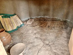 The interior of a model rural traditional hut dwelling in Mbale with earth floor and smooth mud walls. A simple bed made of woven grass mats is laid out on the ground, accompanied by a small grass pillow and a wooden bow. A large clay water pot and a wooden bowl sit in the foreground, while additional mats are leaning against the wall on the left.