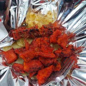 Close-up of grilled red marinated meat served on sliced potatoes in foil, with spices and a fork visible.