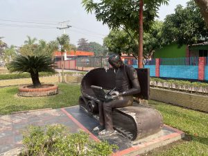 Sculpture of a person sitting on a bench, focused on a laptop. Surrounded by greenery and colorful buildings, conveying a modern, tech-friendly vibe.