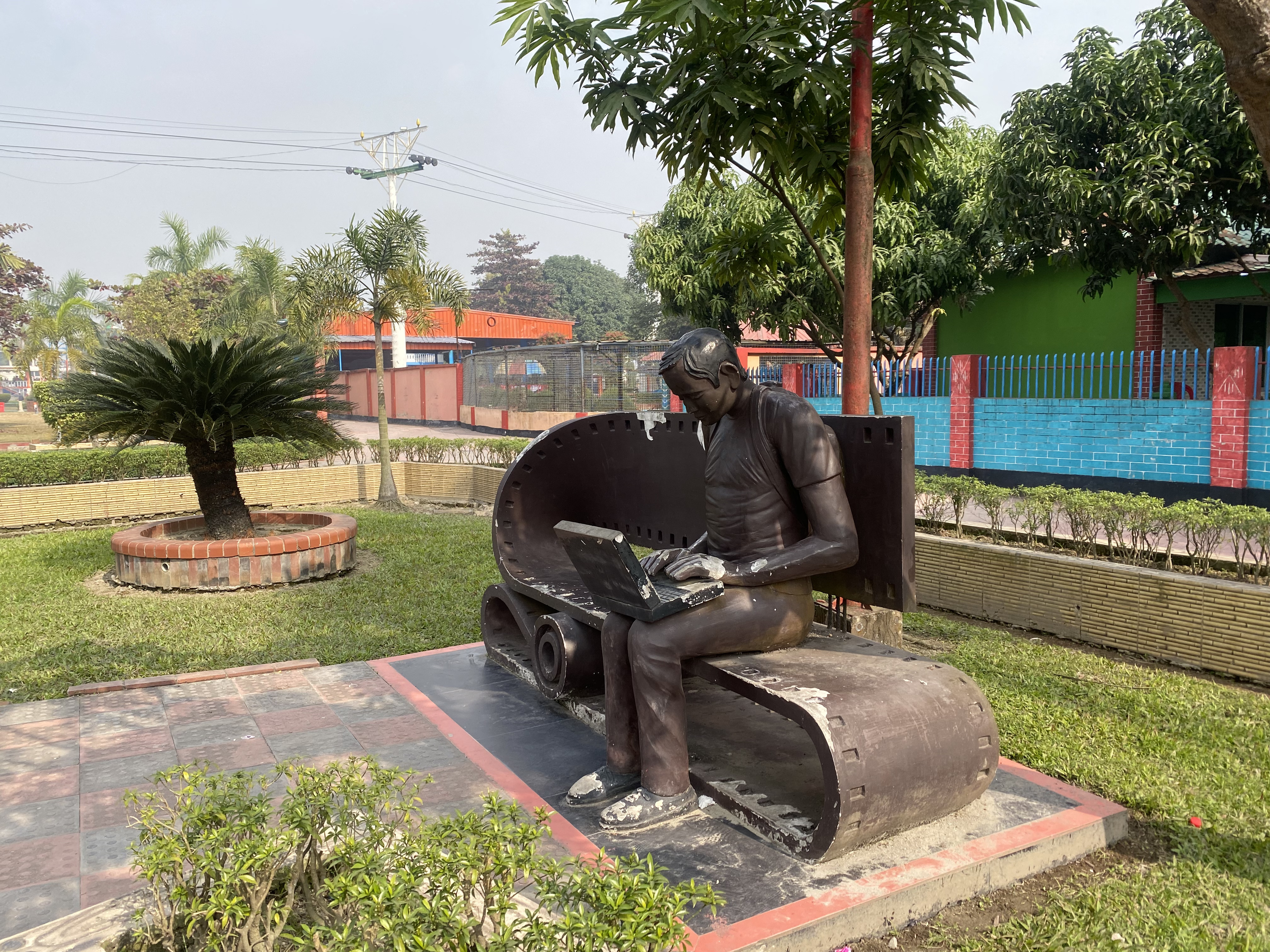 Sculpture of a person sitting on a bench, focused on a laptop. Surrounded by greenery and colorful buildings, conveying a modern, tech-friendly vibe.
