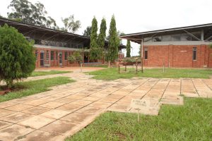 A calm landscaped area with grassy patches and stone pathways leading to modern red-brick buildings with large windows, surrounded by trees including tall slender ones, and a small wooden sign in the foreground reading “Be friendly and approachable,” under an overcast sky.