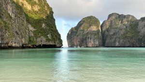 A tropical bay with calm green water, clear pale sand, tree-covered cliffs, sunlit mountains, and small boats in the distance under a bright sky.
