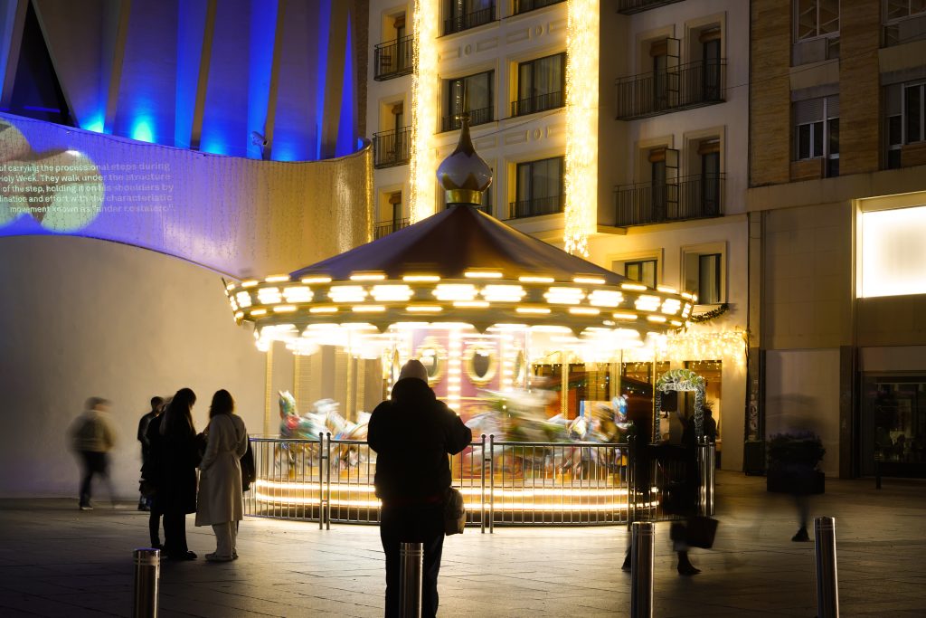 The image depicts a festive carousel illuminated with bright, twinkling lights, spinning merrily in the center. Surrounding the carousel are several people, some standing still and others walking past, creating a lively atmosphere.