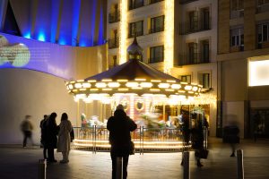 The image depicts a festive carousel illuminated with bright, twinkling lights, spinning merrily in the center. Surrounding the carousel are several people, some standing still and others walking past, creating a lively atmosphere.

