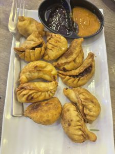 
A neatly arranged plate of golden-brown dumplings, varying in shape and size, is displayed alongside two small bowls of dipping sauces.