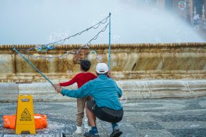 A young child in a red shirt stands in front of an older man, who is crouching down and wearing a blue jacket and black shorts. They are playing with a large bubble wand near a fountain, with bubbles floating around them.