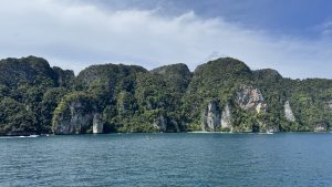 A wide view of a lush, forested tropical island with sheer rock faces and a small white sand beach visible in the distance.