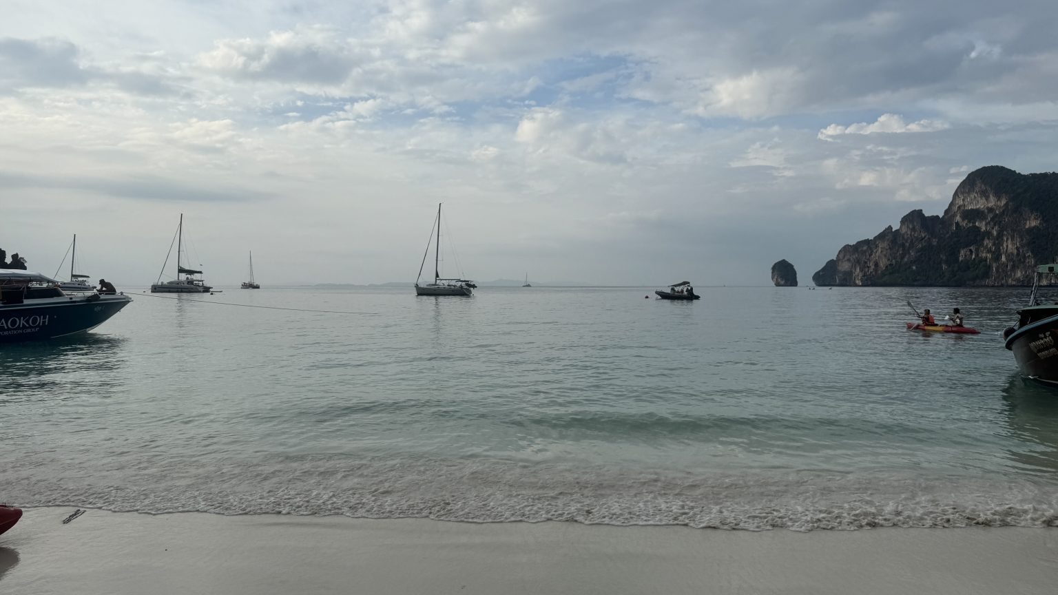 A wide view of a white sand beach with several sailboats and tour boats anchored in the calm, clear turquoise water under a cloudy sky.