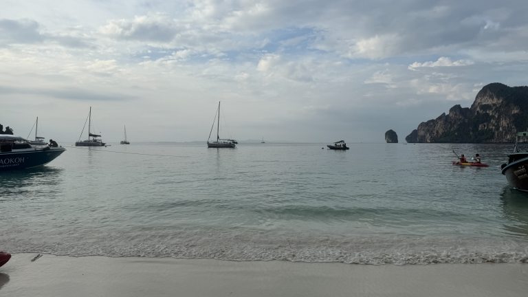 A wide view of a white sand beach with several sailboats and tour boats anchored in the calm, clear turquoise water under a cloudy sky.