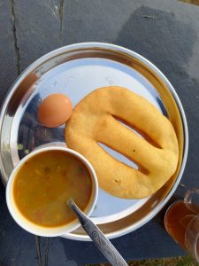 A round stainless steel plate holds a serving of food. To the left, there is a small, brown egg resting on the plate. In the center, a small white bowl contains a yellowish-orange curry with visible bits of vegetables. 