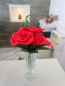 A bouquet of vibrant red paper roses arranged with green leaves in a tall, clear crystal vase. The vase sits on a white counter. In the blurred background, there is a stone-textured wall and a small home shrine.

