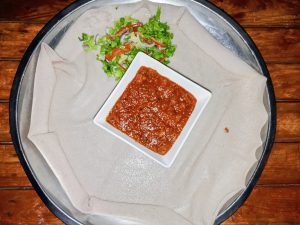 Top-down view of a traditional Eritrean meal with spicy red tibsi in the center, served on injera with a fresh salad on the side.
