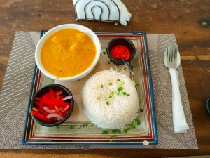 A plated meal with creamy butter chicken in a white bowl, served with steamed white rice topped with green onions. It includes two sides: onion-tomato salad and red chili sauce, served on a square plate over a wooden table.