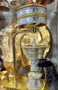 
A metallic decorative object with intricate designs and engravings, featuring a cup-like structure at the bottom with a white wax candle shaped like a teardrop resting inside.