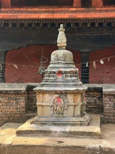 A small, ancient stone stupa, known as a chaitya, sits on a raised platform in a courtyard. The structure features a dome-shaped top and a base with carved niches containing figures of the Buddha. Red tikka powder marks are visible on the carvings. In the background, a traditional brick building with dark, intricately carved wooden beams and a hang