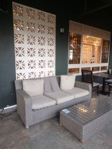 A cozy seating area with a gray wicker sofa and beige cushions beside a glass-top coffee table, set against a decorative white wall with floral cut-out patterns, a large wood-framed window letting in natural light, and a tiled stone floor that enhances the warm, inviting atmosphere.