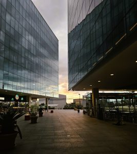 A modern urban setting featuring two tall glass buildings with sleek designs. The space between the buildings reveals a cloudy sky with hints of sunset colors. 
