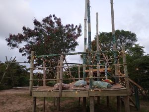A wooden lounge tree-house platform built on stilts and decorated with strings of warm fairy lights. The platform is covered with woven mats and several patterned cushions in shades of green, blue, and orange. Tall wooden poles and leafy trees surround the structure under a cloudy evening sky.