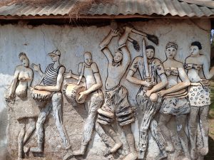 A detailed public bas-relief clay sculpture on an exterior wall in Mbale depicting a line of figures in a traditional celebration. The scene includes drummers, dancers with spears and shields, and women in traditional attire. The figures are rendered in earth tones against a white wall under a rustic thatched and corrugated metal roof.