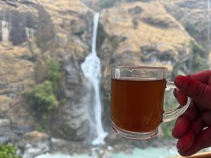 A person holding a glass mug of tea, with a waterfall on a rocky cliff in the background.