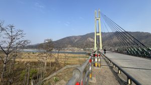 

A scenic view of a suspension Karnali bridge with tall, green towers and supporting cables, set against a backdrop of mountains and a clear blue sky. 