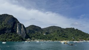 Looking out across a vibrant blue bay at a tropical island coastline featuring steep cliffs and a beach crowded with tour boats.
