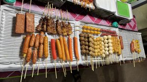 Street food snacks on wooden sticks sit on a metal rack at an outdoor market with a pink-and-white table cover. They include orange hot dogs, red crab sticks, white fish balls, brown meat balls, and grilled meat skewers—some round, some long.
